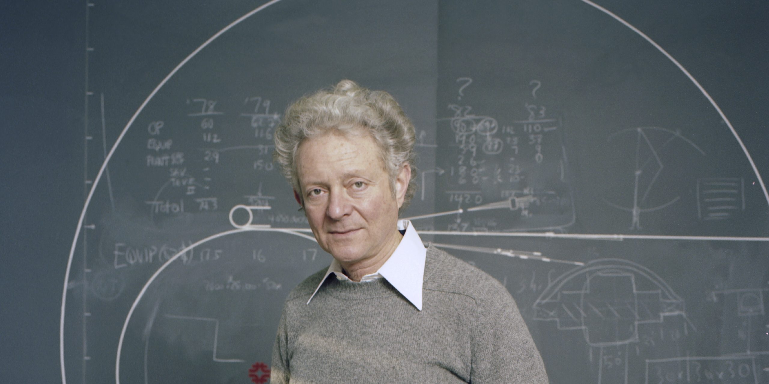 Leon Lederman stands in front of a chalkboard.