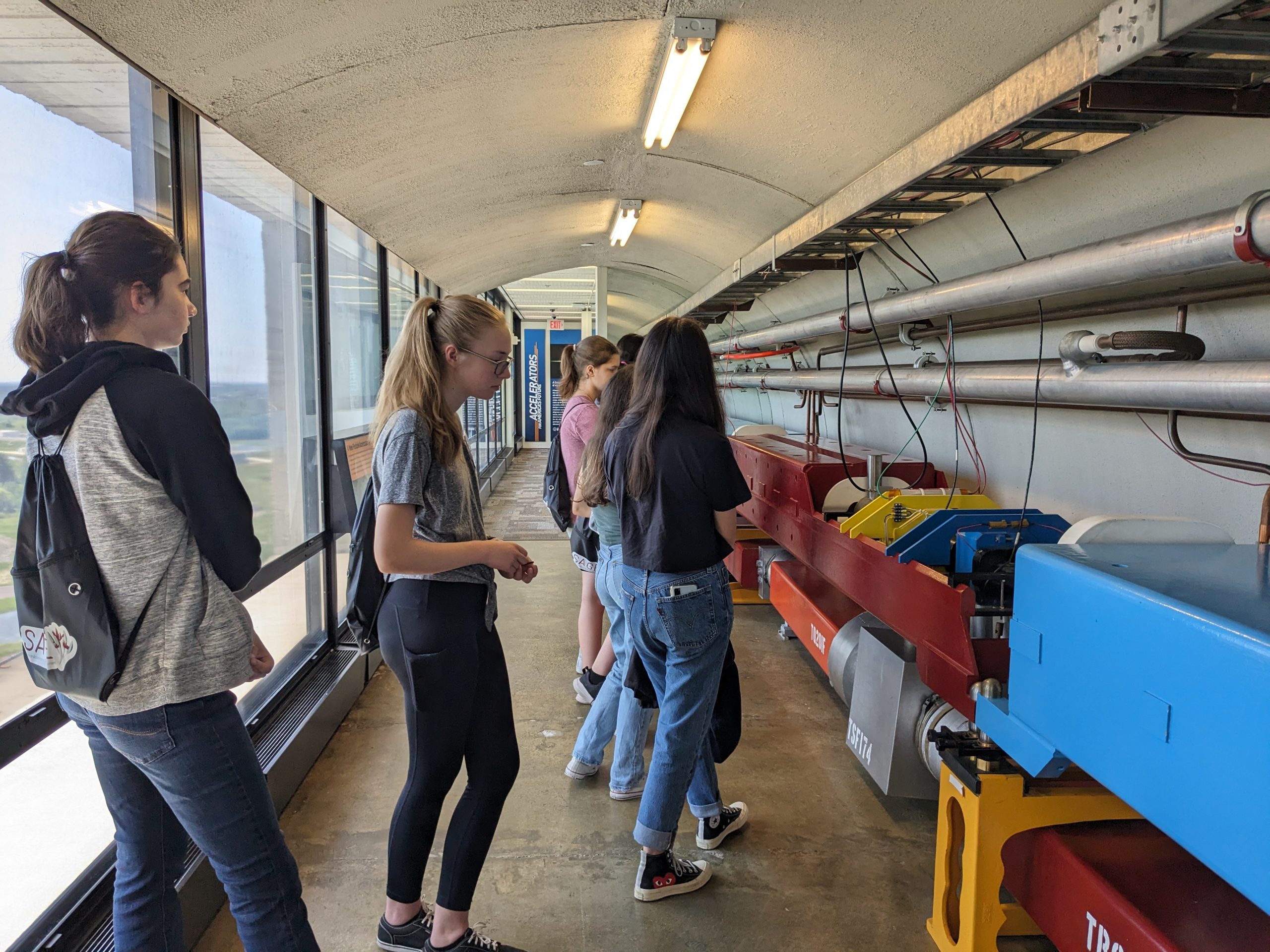 Students stand in a hallway built to mimic the underground tunnels of the accelerator. Pipes and colorful magnets fill the right-hand side. 