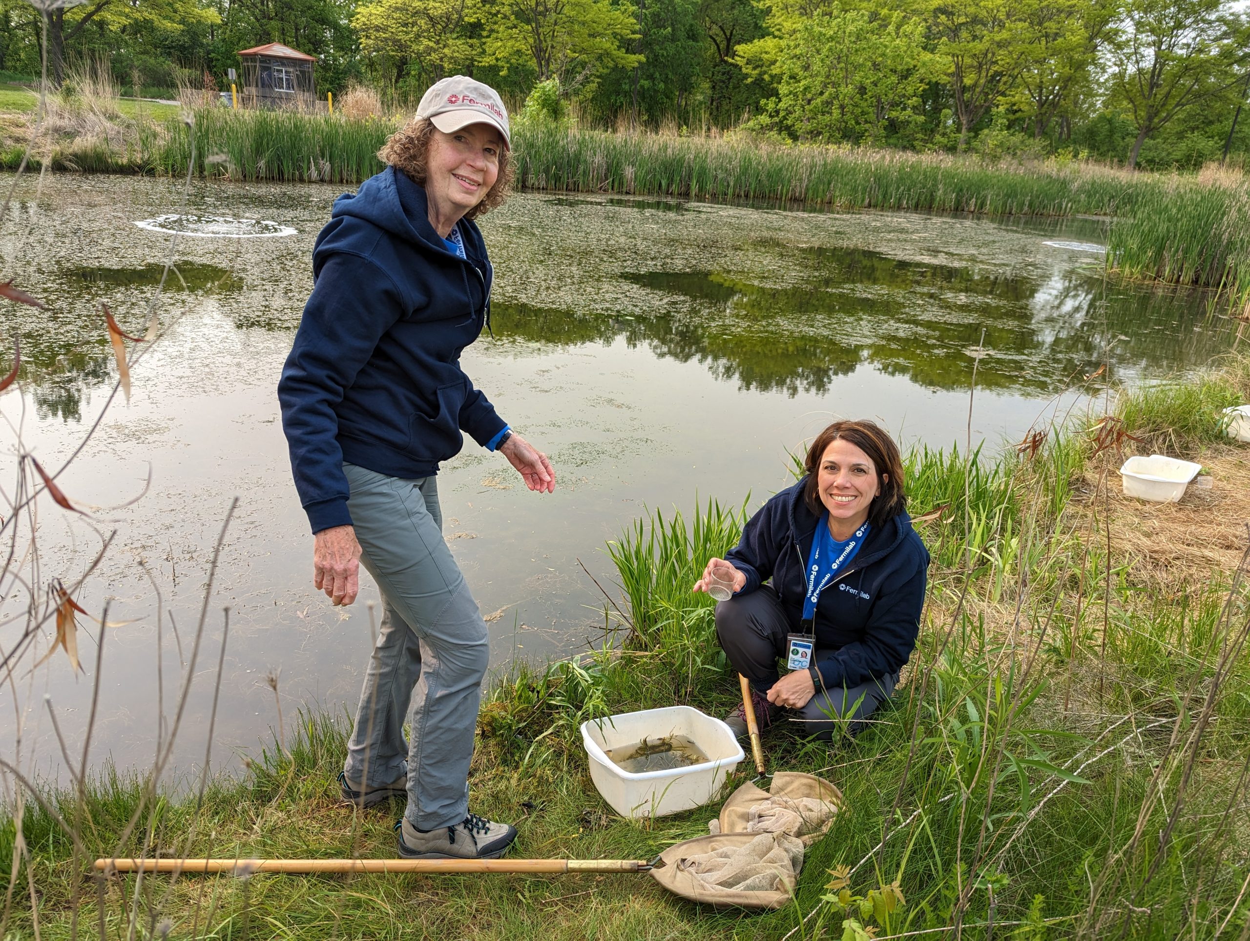 2 Fermilab staff members next to a pond. one is standing and the other is crouched next to a net and bucket. 